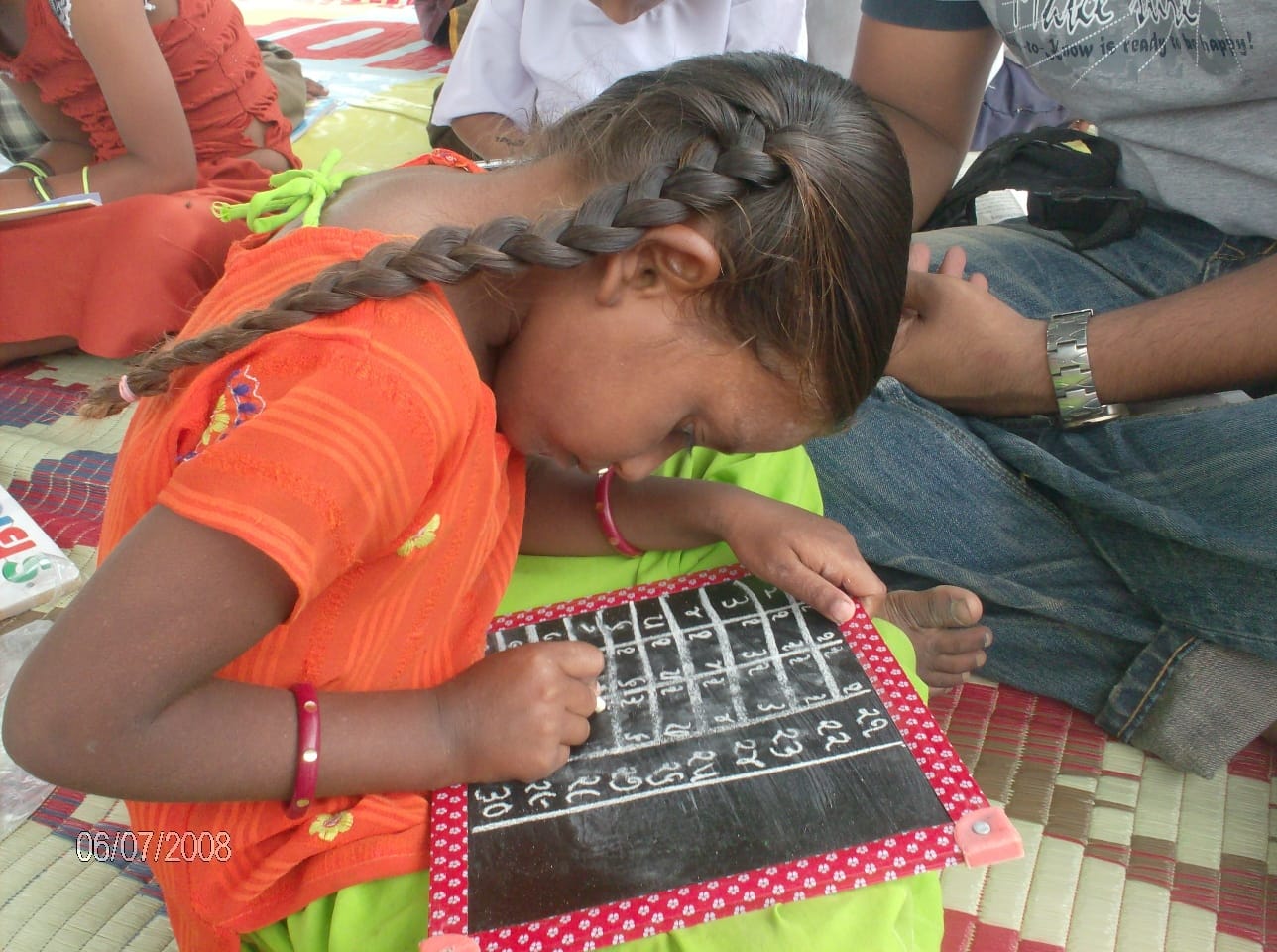 A young girl writes numbers on a slate with other children nearby; the photograph bears the date 06/07/2008.