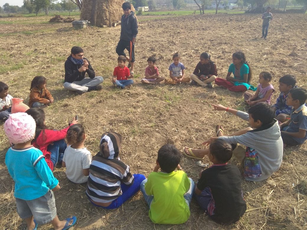 Children sit in a circle in a field with a volunteer facilitating an outdoor session.