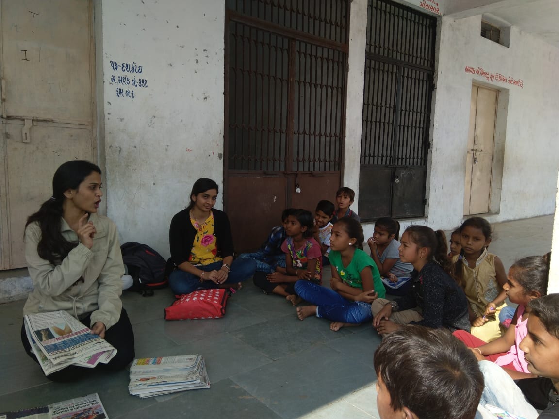 Volunteer/teacher sitting with children outside a community/school building; newspapers/materials visible