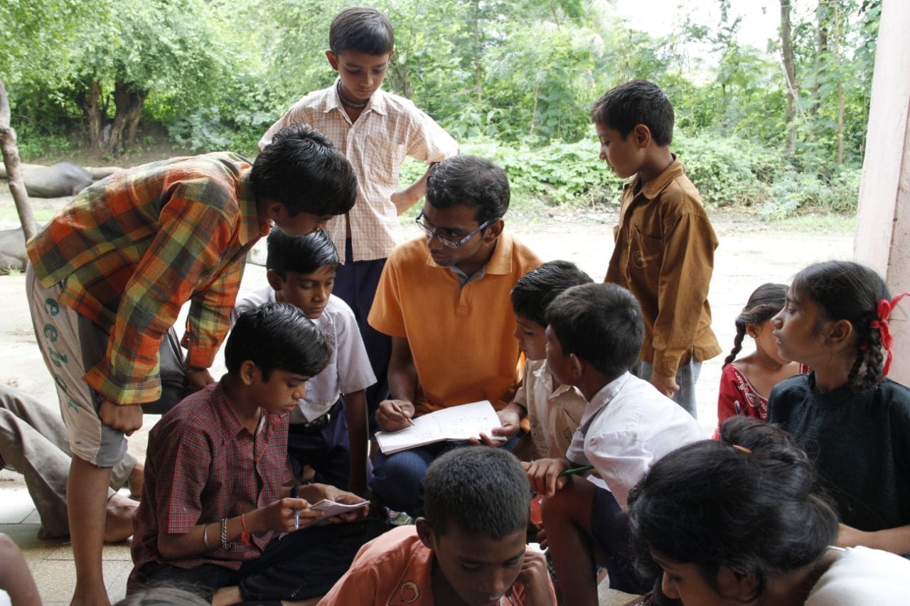 Volunteer sitting among children, looking at their notebook/work while children actively engage