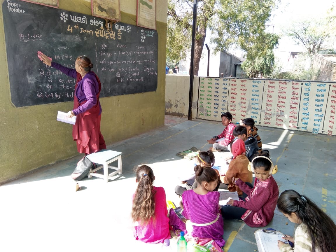 Children learn in an outdoor classroom at Paldi Kankaj, with a volunteer at a blackboard.